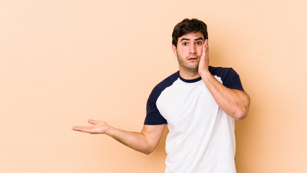 Young man isolated on beige impressed holding copy space on palm.