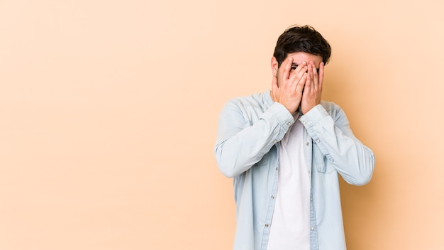 Young man isolated on beige wall blink at the front through fingers, embarrassed covering face