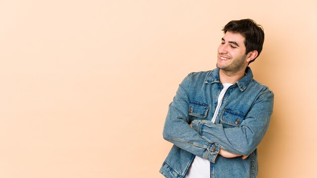 Giovane uomo isolato sul muro beige sorridente fiducioso con le braccia incrociate.