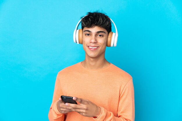 Young man over isolated blue background listening music with a mobile and looking front