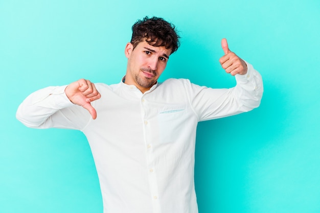 Young man isolated on blue wall showing thumbs up and thumbs down, difficult choose concept