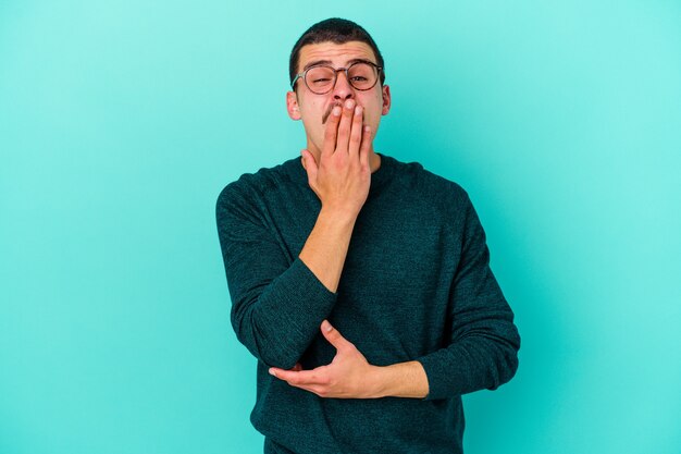 Young man isolated on blue wall yawning showing a tired gesture covering mouth with hand
