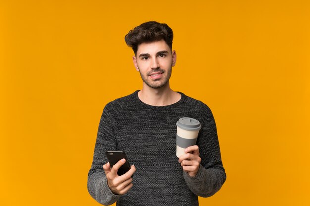 Young man over isolated holding coffee to take away and a mobile