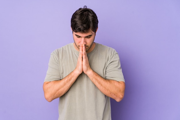 Young man isolated on purple background praying, showing devotion, religious person looking for divine inspiration.