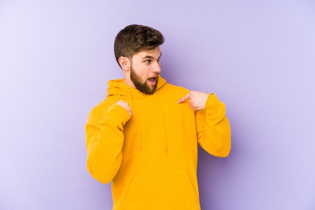 Young man isolated on purple wall surprised pointing with finger, smiling broadly.