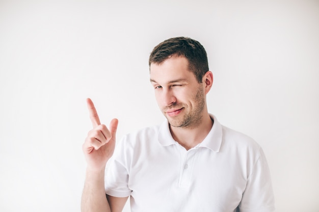 Young man isolated pver white wall. Passionate tricky guy posing alone on camera with finger up. Keep one eye closed.