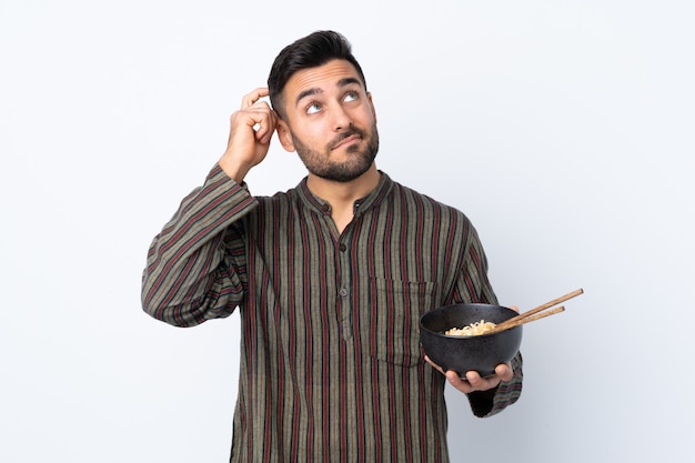 Young man over isolated wall having doubts and with confuse face expression while holding a bowl of noodles with chopsticks