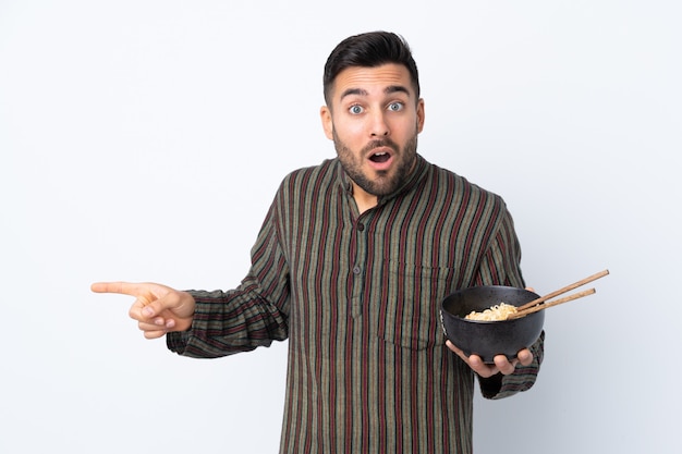 Young man over isolated wall surprised and pointing side while holding a bowl of noodles with chopsticks