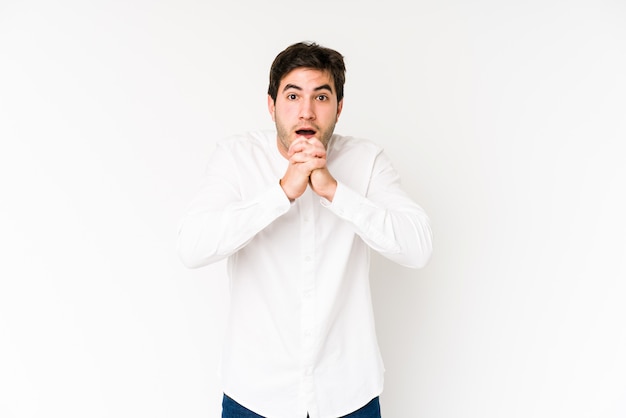 Young man isolated on white praying for luck, amazed and opening mouth looking to front.