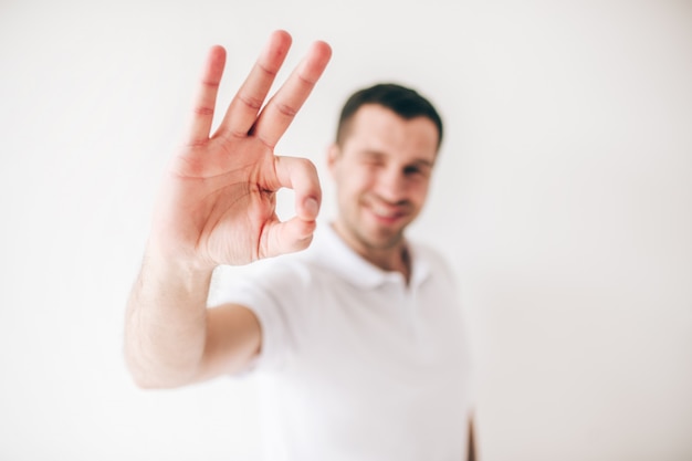 Young man isolated over white wall. Cheerful positive guy on blurred show ok symbol with fingers. Satisfied person.