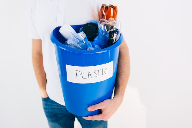 Young man isolated over white wall. Cut view of guy holding blue bucket with plastic inside it. Recycling time for no waste lifestyle. Good for environment.