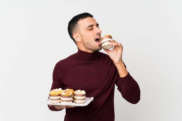 Young man over isolated white wall holding mini cakes and eating it