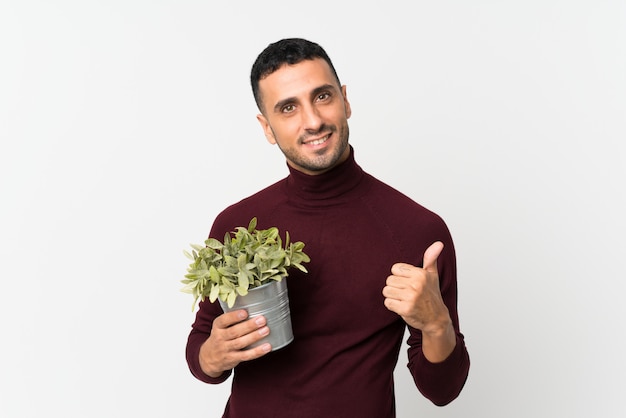 Young man over isolated white wall taking a flowerpot and with thumb up