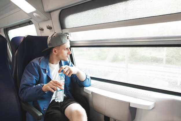 Young man in a jacket and cap sits in a train with a bottle of water in his hands and looks window.