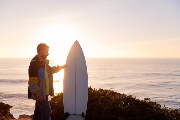 Young man in jacket holding his surfboard while looking at the sunset by the sea leisure and hobbies concept copy space for text