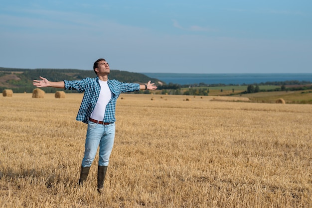 Молодой человек в джинсах, рубашке, резиновые сапоги в поле с открытыми руками