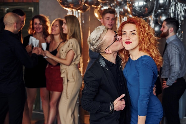 Young man kisses redhead woman against their friends in christmas decorated room and celebrating New Year.