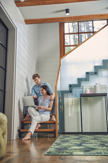 young man and lady sitting on the stairs in luxury modern house and watching to the screen computer