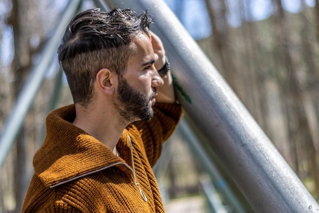 A young man leaning on a metal structure in the open air looking away