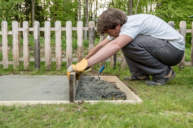 Young man leveling cement in a backyard using wooden plank laying new concrete foundation in a diy concept.