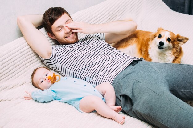 若い男は子供と彼の犬と一緒にベッドに横たわっています。笑顔、幸せ、笑い、休息。子供とペットのコンセプト