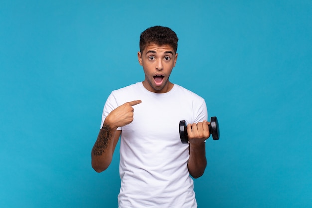 Young man lifting a sumbbell feeling happy, surprised and proud, pointing to self with an excited, amazed look