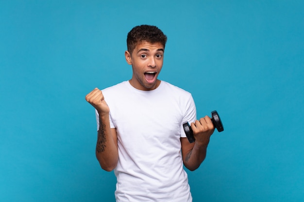 Young man lifting a sumbbell feeling shocked, excited and happy, laughing and celebrating success, saying wow!