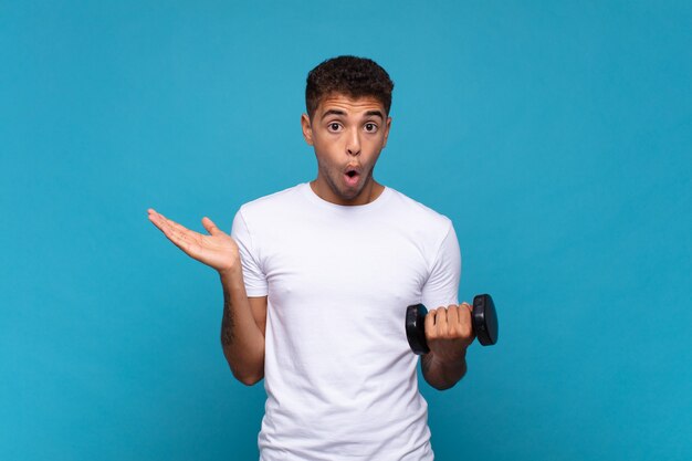 Young man lifting a sumbbell looking surprised and shocked, with jaw dropped holding an object with an open hand on the side