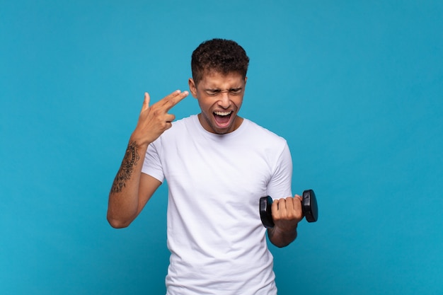 Young man lifting a sumbbell looking unhappy and stressed, suicide gesture making gun sign with hand, pointing to head