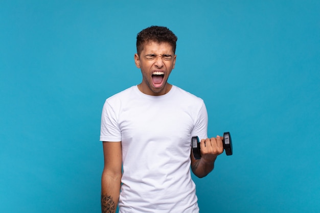 Young man lifting a sumbbell shouting aggressively, looking very angry, frustrated, outraged or annoyed, screaming no