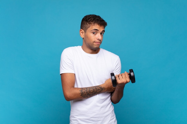 Young man lifting a sumbbell shrugging, feeling confused and uncertain, doubting with arms crossed and puzzled look