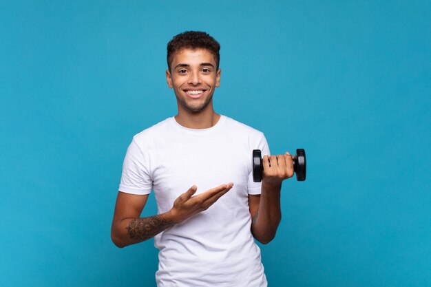 Young man lifting a sumbbell smiling cheerfully, feeling happy and showing a concept in copy space with palm of hand