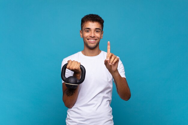 Young man lifting a sumbbell smiling and looking friendly, showing number one or first with hand forward, counting down