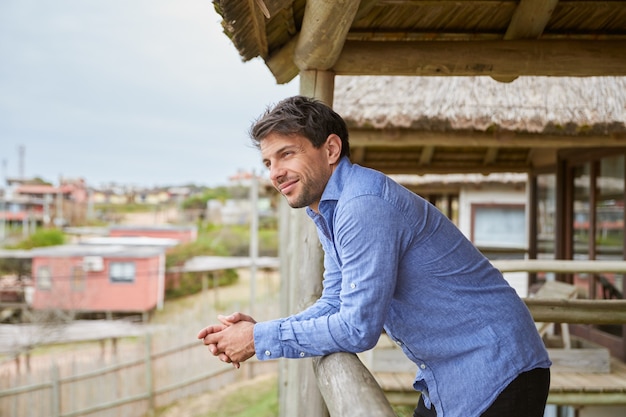 Giovane in camicia azzurra appoggiato alla ringhiera di legno della terrazza della sua cabina mentre sorride.