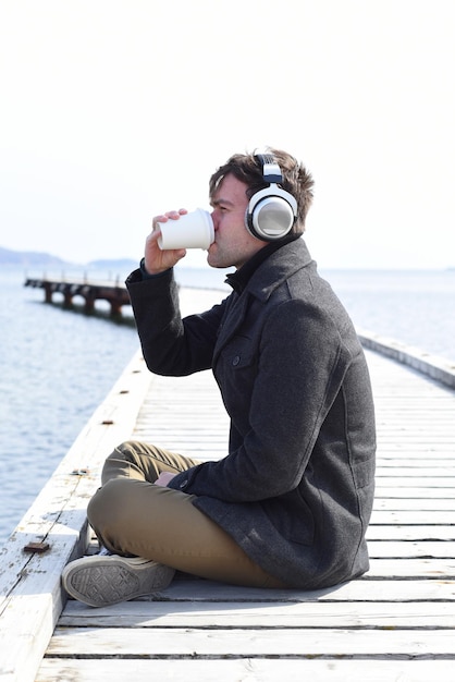 晴れた日に音楽を聴き、桟橋に一人で座ってコーヒーを飲む若い男