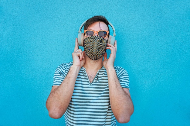 Young man listening music with big headphones and face mask on\
in coronavirus time boy in summer bright background feeling\
cheerful in social distance with sunglasses