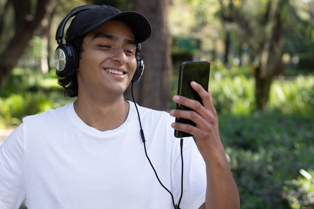 Young man listening to music with headphones and looking at his cellphone screen while strolling through the park smiling and relaxed in the middle of nature