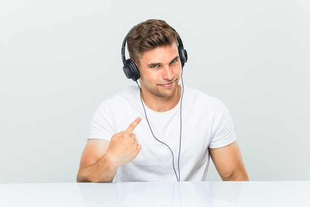 Young man listening to music with headphones pointing with finger at you as if inviting come closer.