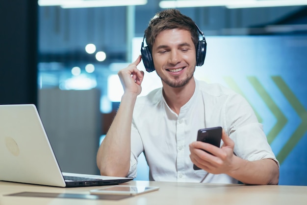 A young man listens to podcasts in headphones holds a mobile phone in his hands talks on video communication Sitting at work in the office with a laptop