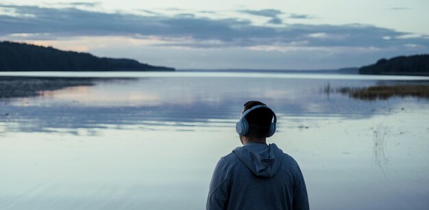 若い男が夕方に湖の近くでヘッドフォンでリラックスした音楽を聴きます 自然の中で禅の穏やかさを捉えます