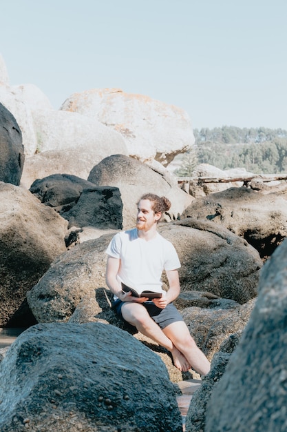 Фото Молодой человек смотрит в сторону от камеры, читая книгу на берегу моря, копируя пространство, уверенность и изучая концепции, будущее и самопомощь