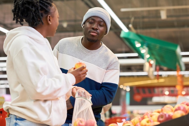 Young man looking at fresh apple held by his girlfriend in supermarket