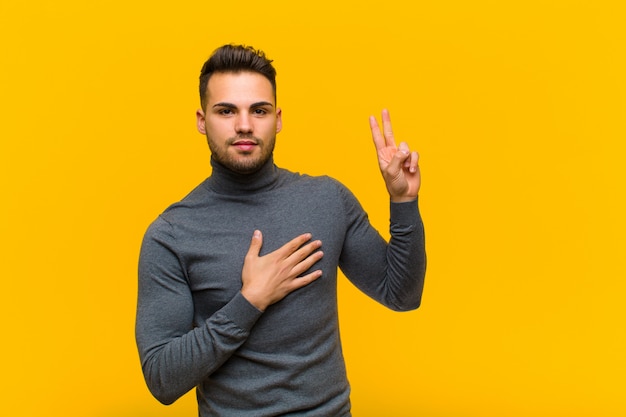 Young man looking happy, confident and trustworthy, smiling and showing victory sign, with a positive attitude over orange wall