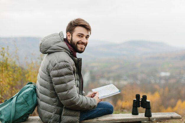 Молодой человек смотрит на карту, сидя на деревянной скамейке в горах