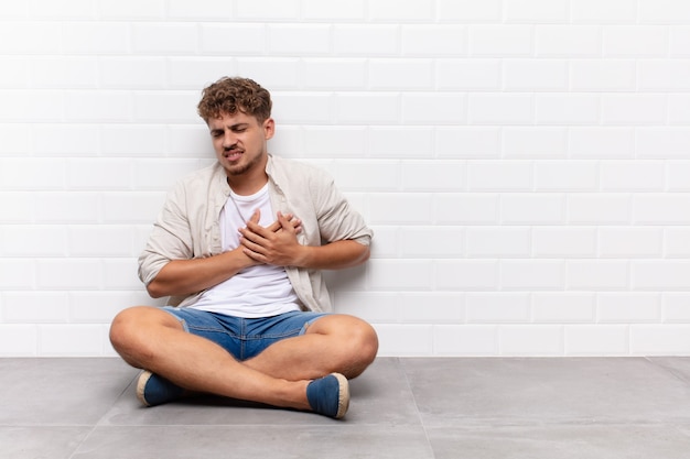 Giovane che sembra triste, ferito e affranto, tenendo entrambe le mani vicino al cuore, piangendo e sentendosi depresso