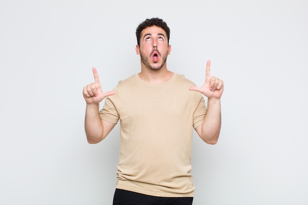 Young man looking shocked, amazed and open mouthed, pointing upwards with both hands to copy space