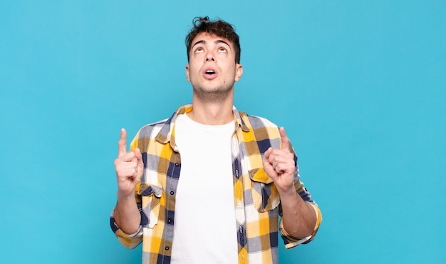 Young man looking shocked, amazed and open mouthed, pointing upwards with both hands to copy space