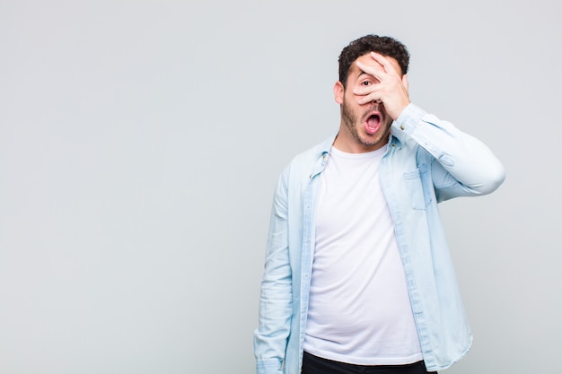 Young man looking shocked, scared or terrified, covering face with hand and peeking between fingers