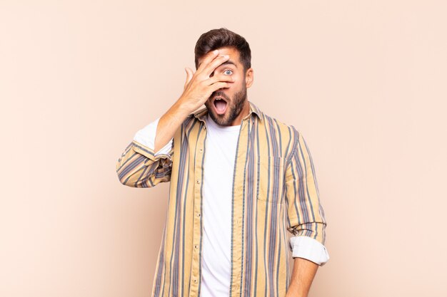 Young man looking shocked, scared or terrified, covering face with hand and peeking between fingers