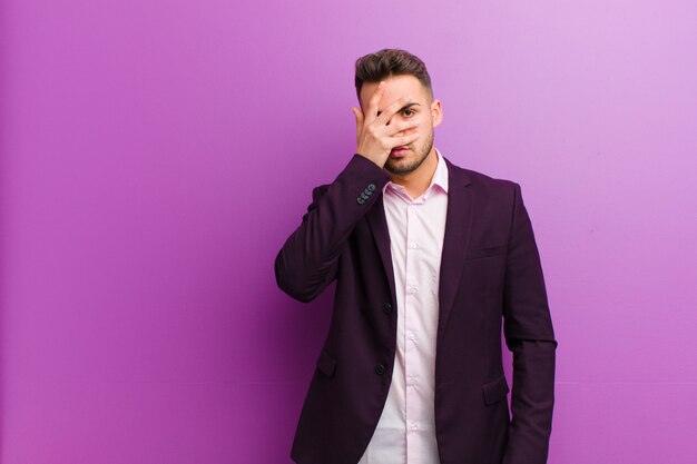 Young man looking shocked, scared or terrified, covering face with hand and peeking between fingers